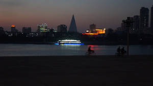 單車手在黃昏時沿河岸騎行，背景有燈火通明的遊輪和城市天際線。