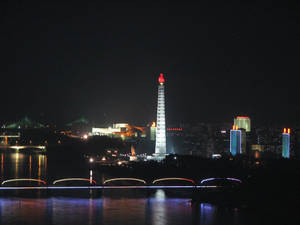 夜間景色，一座發光的紀念碑立於水上，背景有橋樑和城市燈光。