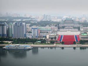 平壤城市景觀的航拍圖，河岸上有大型旗幟排列和水上的船。 — 韓國 壁紙