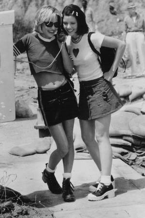 兩位穿著90年代服裝的女性，笑著共享一部有線電話在戶外。