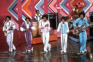 Jackson Five 在帶有幾何圖案和反射地板的色彩繽紛舞台上表演。 — 米高積遜 GIF 壁紙