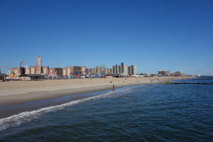 海浪拍打沙灘，背景有遊樂園和建築物，天空清澈湛藍。