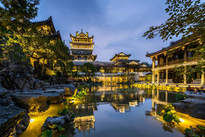 古老的中式亭閣在黃昏時亮起，倒映在寧靜的池塘中，周圍有岩石和樹木。 — 唐人街 GIF 壁紙