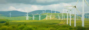 一排排風力發電機矗立在鬱鬱蔥蔥的綠色田野中，背景有山丘和多雲的天空。 — 超寬螢幕 壁紙