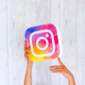 雙手拿著水彩Instagram標誌毛絨玩具，背景是白色木頭。