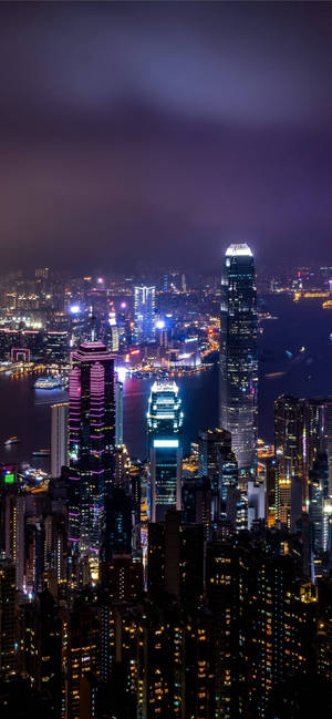 密集的城市天際線夜景，閃爍的摩天大樓和海濱燈光。 — iPhone 15 Plus 壁紙