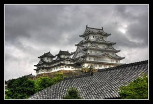 山頂上的白色日本城堡，深色瓦片屋頂和綠樹，多雲天空。