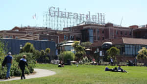 人們在陽光明媚的城市公園中，在 Ghirardelli 招牌附近的草地上放鬆。 — 花園牆外 壁紙