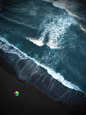 從高空拍攝，海浪拍打黑色沙灘，有一把細小的彩色雨傘。 — 海浪 壁紙