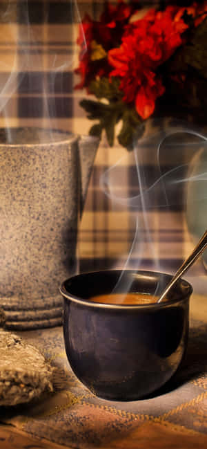 冒煙的咖啡杯和陶瓷杯放在附近帶有紅花的格紋布上。