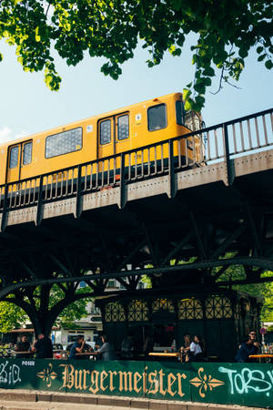 高架軌道上的黃色火車，下方是熙熙攘攘的戶外咖啡館，有人群和綠樹。