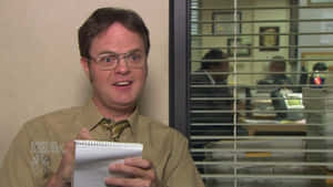 《辦公室》的 Dwight Schrute 在辦公室拿著筆記本微笑。