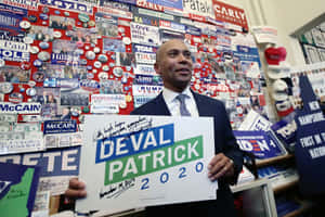 男子舉起 Deval Patrick 2020 競選標語牌，周圍環繞著政治紀念品。
