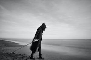 一個戴著兜帽的人在海灘上手持長杖，暮色中霧氣瀰漫。 — 黑白 壁紙