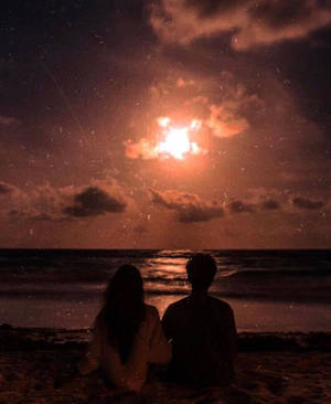 兩個人夜晚坐在沙灘上，透過雲層觀看月亮。