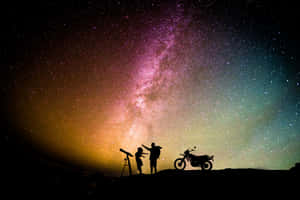 兩個人和一輛電單車以色彩繽紛的星空和銀河為背景形成剪影。