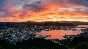 日落時分的沿海城市，橙色和粉紅色的天空倒映在水面和背景的山巒上。