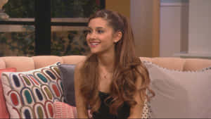 Ariana Grande 在訪問期間坐在粉紅色沙發上，與圖案靠墊一起微笑。