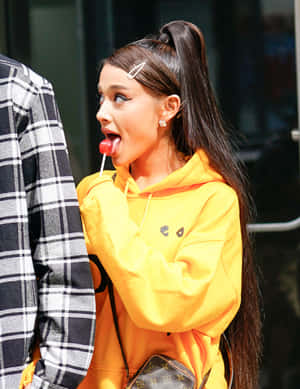 Ariana Grande 身穿黃色連帽衫，在戶外頑皮地舔著紅色棒棒糖。