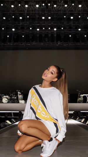 Ariana Grande 身穿白色Reebok服裝，在明亮的燈光下蹲在音樂會舞台上。