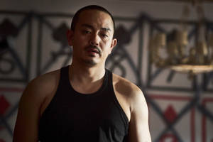 一個穿著黑色背心的男人在一個有裝飾性牆壁和吊燈的房間裡嚴肅地凝視。