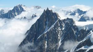 一座高聳的山峰在翻騰的雲層之上崛起，積雪附著在岩石斜坡上。