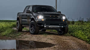 黑色福特 F-150 Raptor 貨車喺泥濘小徑上，喺暴風雨天空下嘅水氹倒影。
