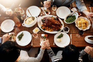 Family gathers around a wooden table for a holiday meal with hands clasped in prayer.