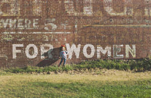 一個戴著紅色蝴蝶結的小女孩站在一面磚牆前，牆上有褪色的「FOR WOMEN」塗鴉。