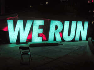 夜間以紅色背光照明的巨大發光青綠色字母WE RUN，附近有一張椅子。 — 跑步 壁紙