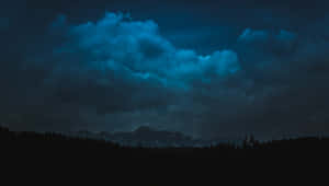 在發光的藍色夜空下，帶有戲劇性雲層的黑暗森林和山脈。 — 夜空 壁紙