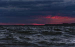 暴風雨黃昏天空下，帶有紅雲和遠處陸地的黑暗海浪。 — 海 壁紙