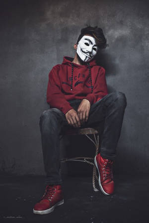 身穿紅色連帽衫和運動鞋，戴著白色 Guy Fawkes 面具的人坐在凳子上。