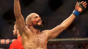 一位滿臉鮮血的MMA拳手在格鬥籠內高舉雙臂慶祝勝利。