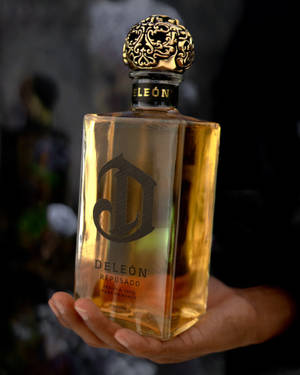 手拿著一個金色的Deleón Reposado龍舌蘭酒瓶，帶有華麗的瓶蓋。 — 4:5 壁紙