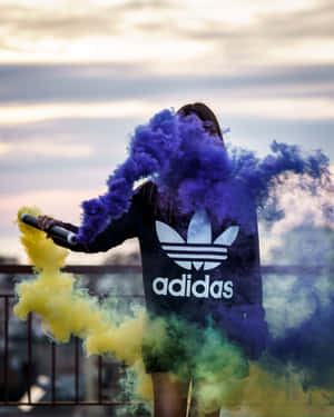 穿著黑色Adidas連帽衫的人在暮色中的屋頂上釋放紫色和黃色煙霧。 — Adidas GIF 壁紙