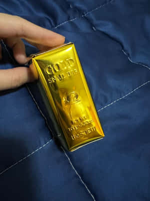 手拿著一塊標有 'GOLD SNAIL BAR' 的金條，放在藍色絎縫布料上。