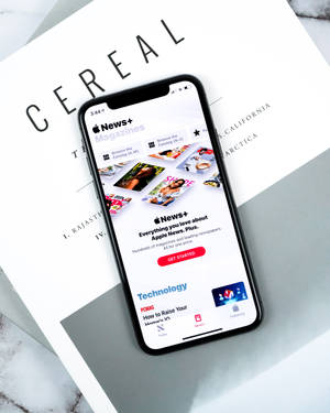 智能手機在大理石表面上顯示 Apple News+ 應用程式，旁邊有一本白色書本名為 CEREAL。