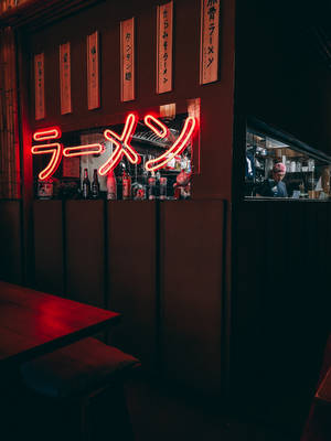 在昏暗的日本餐廳中，發光的紅色拉麵霓虹燈招牌，櫃檯後方有廚師。