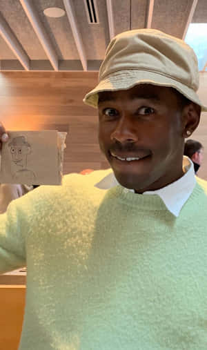 身穿綠色毛衣、戴著米色帽子的男子舉起一張鉛筆素描人臉。 — Tyler the creator GIF 壁紙