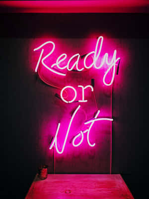 粉紅色霓虹燈牌，上面寫着「Ready or Not」，位於黑暗的牆壁上，下方有一張木桌。 — 霓虹 壁紙