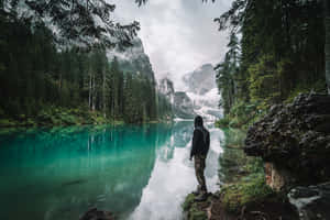 穿著連帽衫的人站在碧綠的湖邊，背景是森林和山巒。