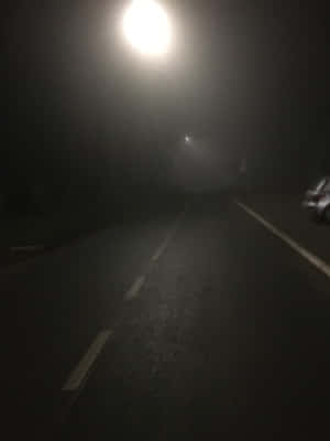 夜晚一條黑暗霧濛濛的道路，由一盞街燈照亮，旁邊可見一輛汽車。