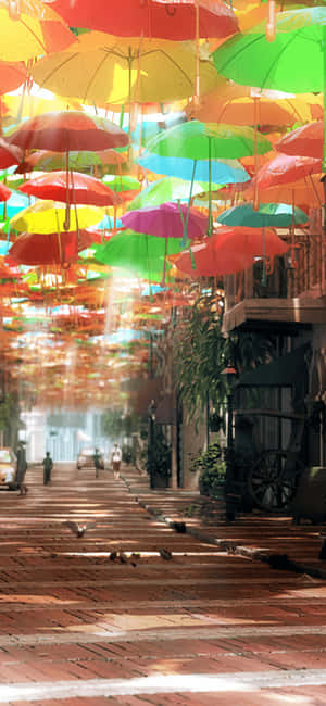 五彩繽紛的雨傘懸掛在頭頂，在有行人的磚砌街道上投下斑駁的光影。 — POCO F7 Pro 壁紙