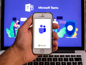手裡拿著手機上的 Microsoft Teams 應用程式，筆記型電腦螢幕顯示應用程式介面。