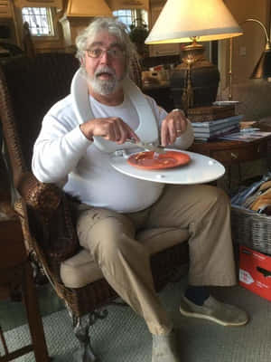 一位戴眼鏡、留白鬍子的老人坐在室內藤椅上，從盤子裡進食。
