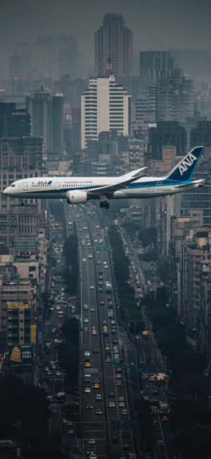 全日空飛機低飛過一條高樓林立、交通繁忙的城市街道。 — Pixel 10 Pro XL 壁紙