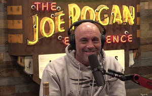 Joe Rogan 喺podcast錄音室入面微笑，身後有霓虹燈牌。
