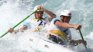 兩名皮划艇運動員在洶湧的白水中航行，表情專注，手持綠色槳。