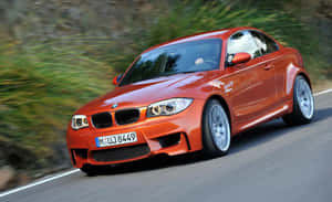 橙色BMW M1雙門跑車在彎曲的道路上疾馳，兩旁是綠色的山丘。